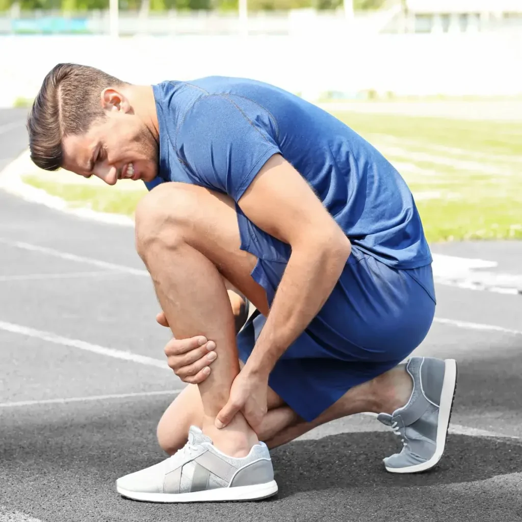 A man in athletic wear is sitting on a running track, holding his ankle in pain as if he is injured. He is wearing a blue shirt, blue shorts, and gray sneakers.