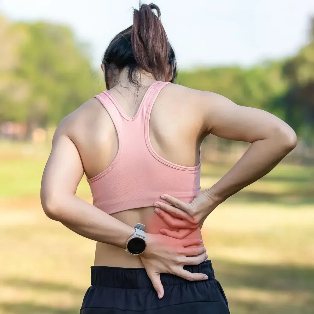 A woman in athletic wear stands outdoors, facing away, holding her lower back with one hand. A red area highlights back pain or discomfort. She appears to be experiencing back pain.