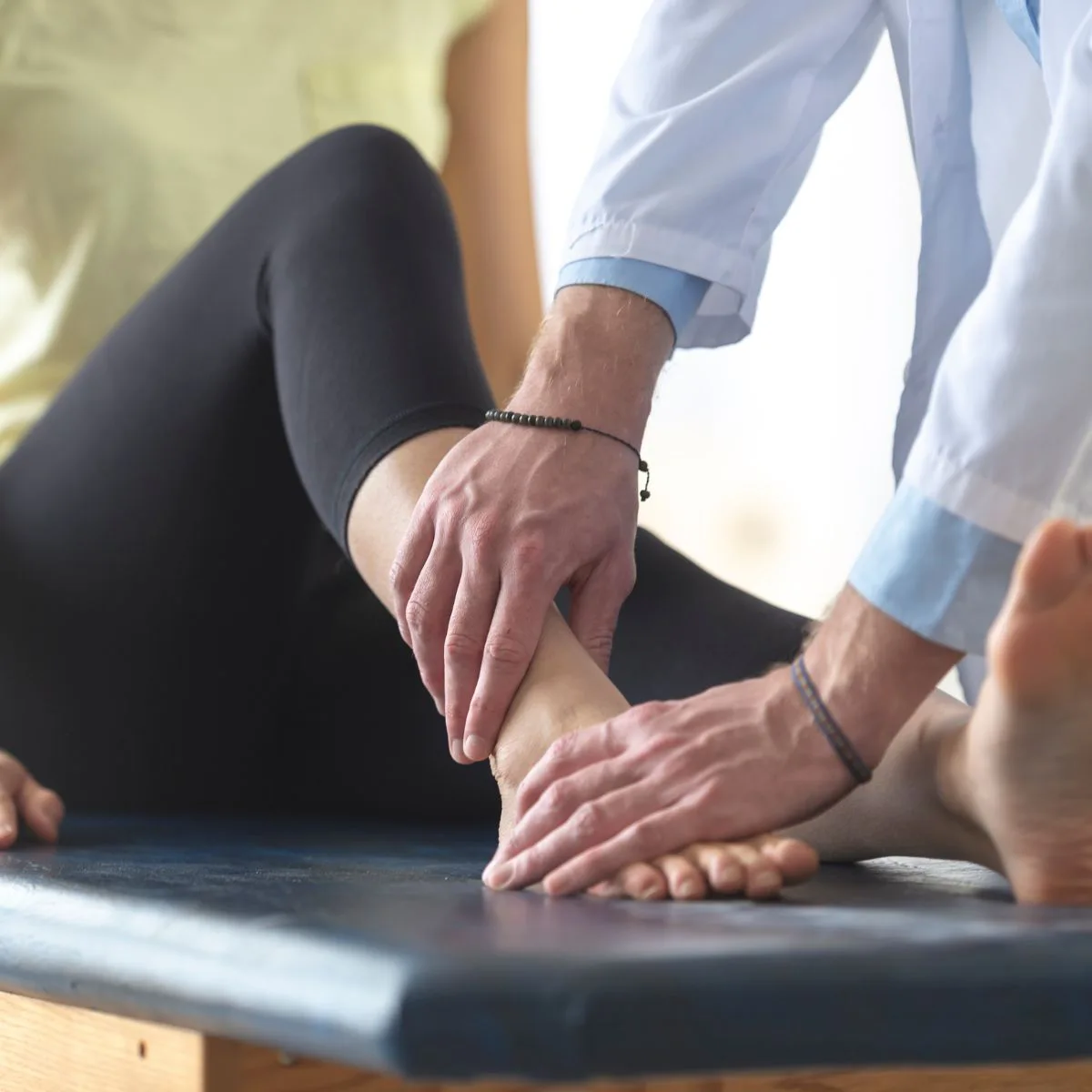 A healthcare professional examines a persons ankle on an examination table. The patient is lying down with their leg extended while the professional gently holds and inspects the ankle.