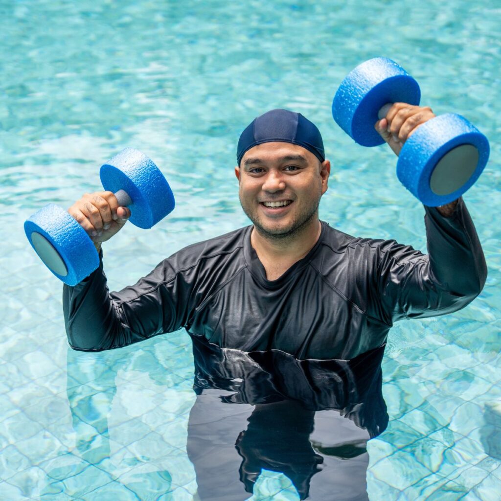 A smiling man in a swim cap and black swim shirt stands in a pool, holding blue foam dumbbells above the water with both hands.