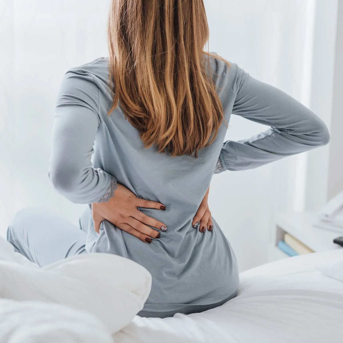 A woman with long hair, dressed in light blue pajamas, sits on a bed with her back to the camera, holding her lower back with both hands as if experiencing pain or discomfort.