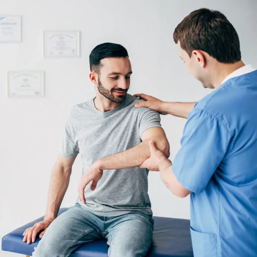 A male physical therapist in blue scrubs examines the arm of a seated man in a gray t-shirt and jeans, gently rotating his elbow in a medical office with certificates on the wall.