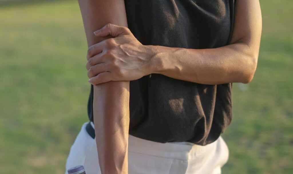 A person standing outdoors holds their right forearm with their left hand, wearing a black sleeveless top and white pants, with a blurred grassy background.