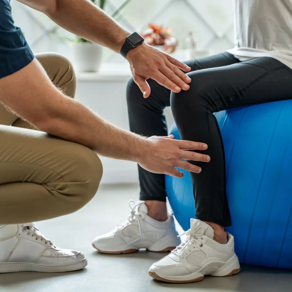 A physical therapist examines a persons knee as they sit on a blue exercise ball. Both are wearing athletic clothes and white sneakers in a bright indoor setting.