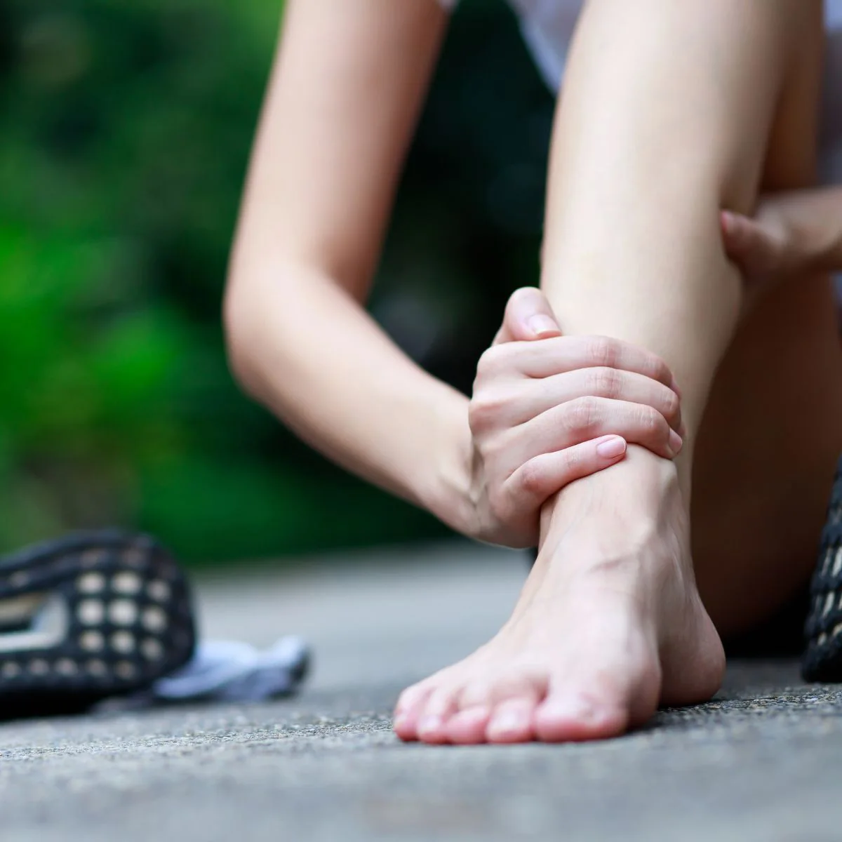 A person sitting on the ground outdoors, holding their ankle with both hands, suggesting pain or injury. One shoe is off and visible next to their bare foot.