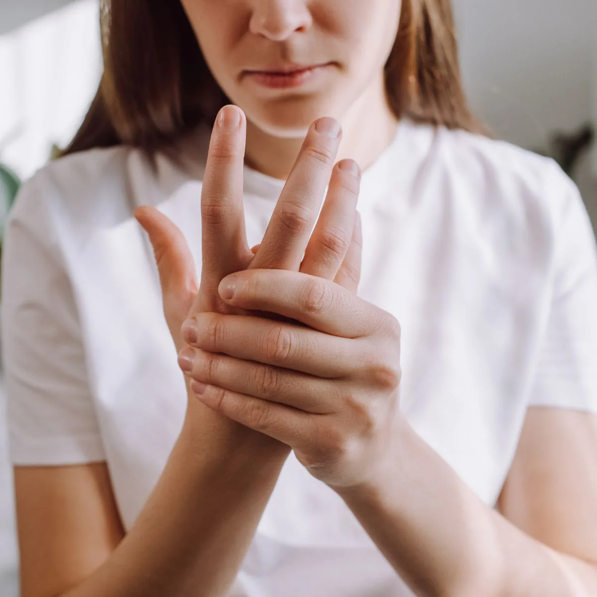 A woman in a white shirt holds and massages her aching hand, appearing to experience pain or discomfort in her fingers.