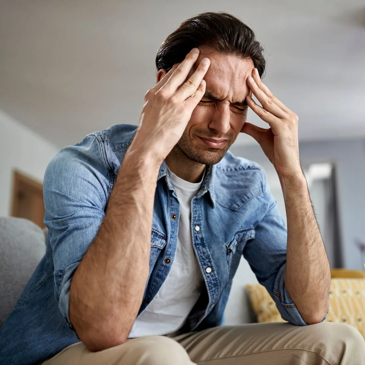 A man sitting on a couch holds his head with both hands and closes his eyes, appearing to be in pain or experiencing a headache. He is wearing a denim shirt and looks distressed.