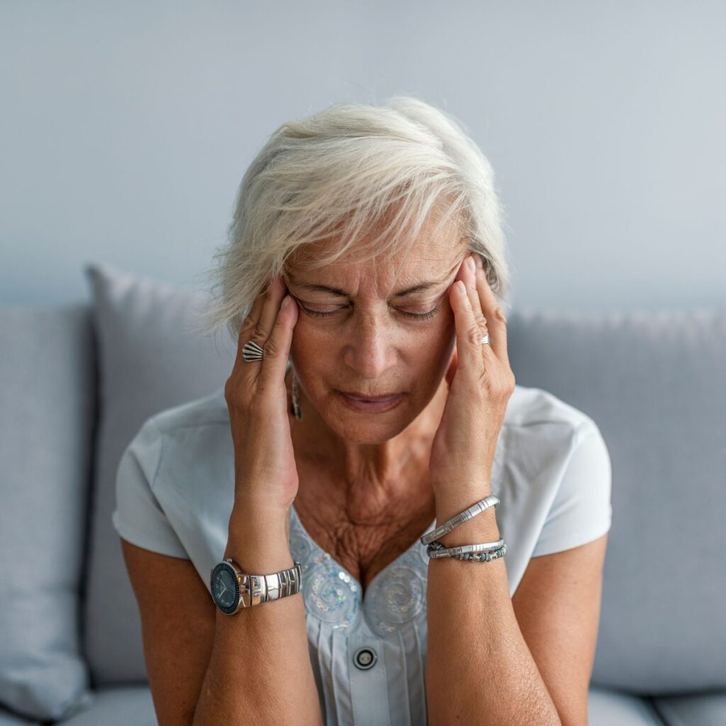 Older woman with short gray hair sits on a couch, eyes closed, pressing her temples with both hands as if experiencing a headache or stress. She wears a light blouse, watch, and bracelets. The background is softly blurred.