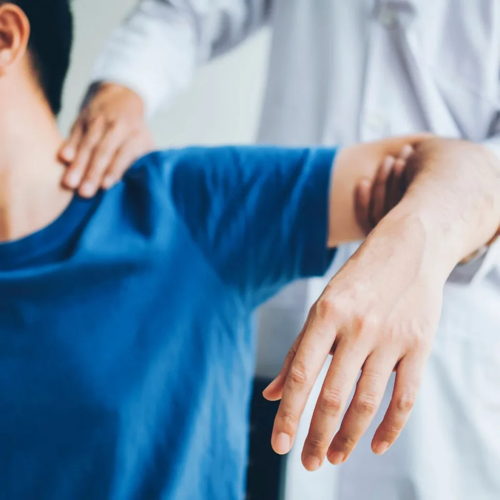 A healthcare professional in a white coat assists a person in a blue shirt by gently stretching their shoulder and arm during a physical therapy or rehabilitation session.