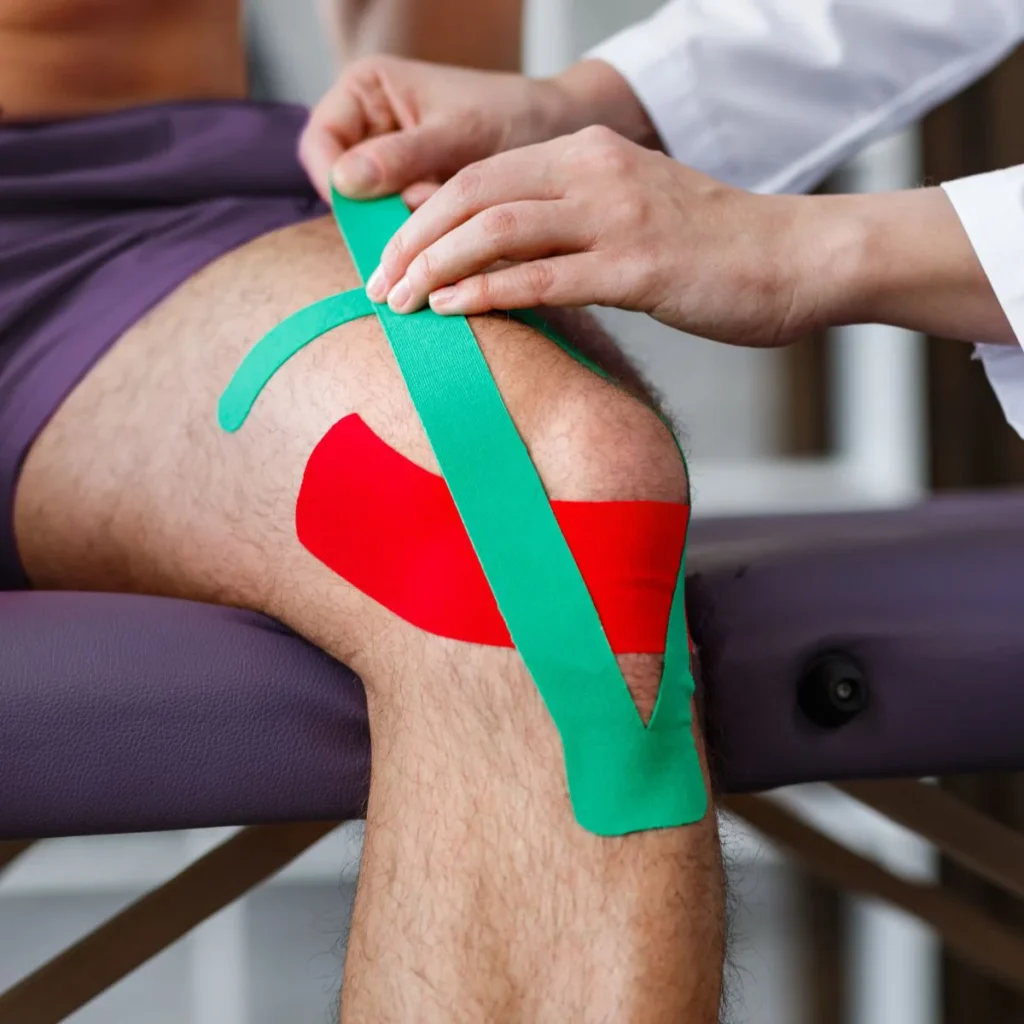A person sits on an exam table while a healthcare professional applies green and red kinesiology tape to their knee.