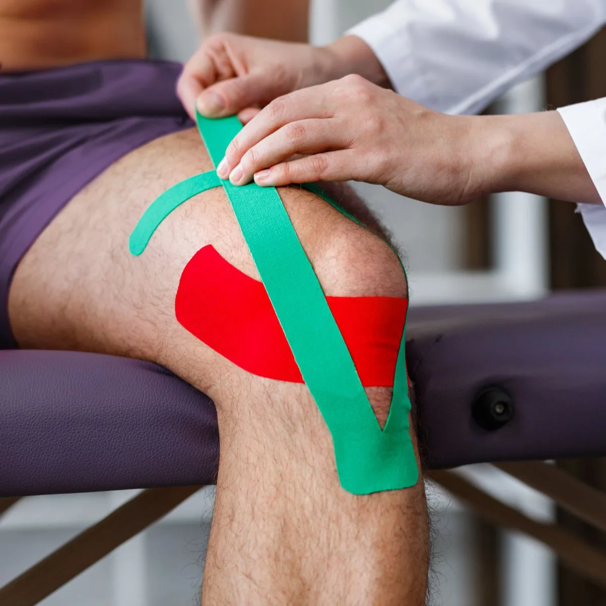 A person sits on an exam table while a healthcare professional applies green and red kinesiology tape to their knee.