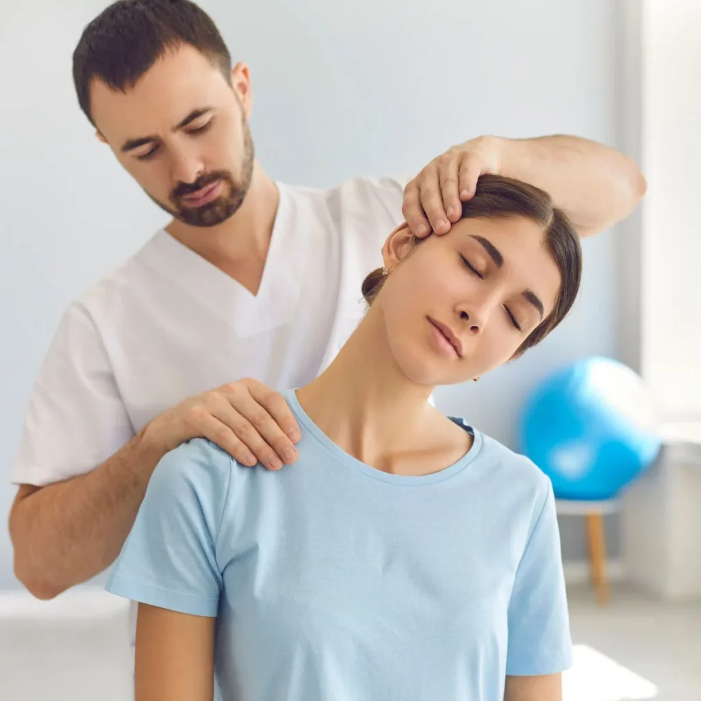 A physical therapist in a white uniform gently stretches a young womans neck while she sits with her eyes closed, wearing a light blue shirt in a bright, modern clinic.