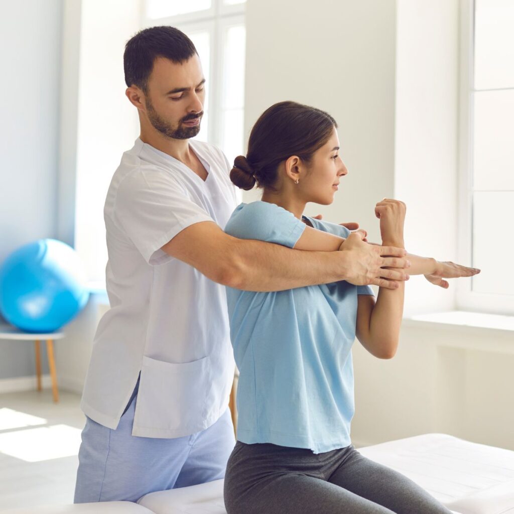 A physical therapist helps a woman stretch her arm in a bright clinic room. The woman sits on an exam table while the therapist gently supports her back and arm. Exercise equipment is visible in the background.