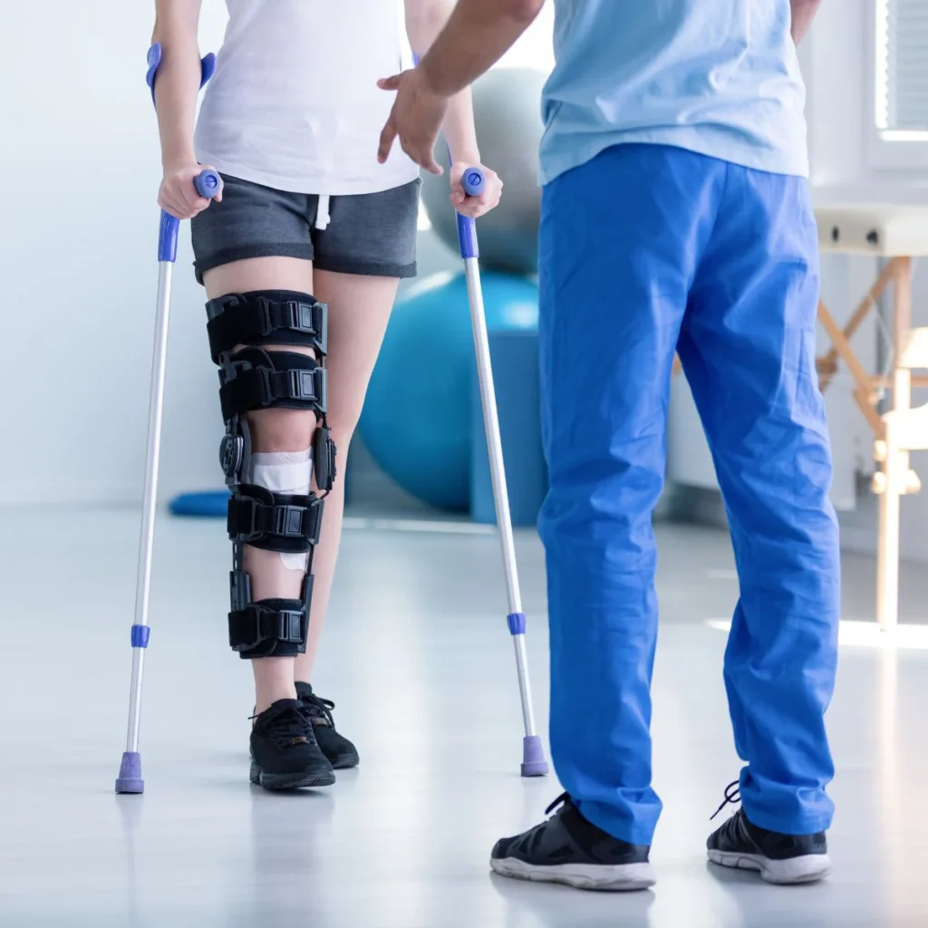 A person using crutches and wearing a black knee brace listens to a healthcare professional in blue scrubs during a physical therapy session.