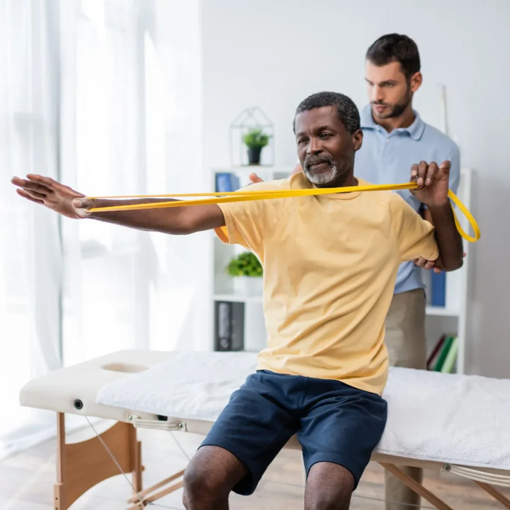 A man sits on an examination table doing a resistance band arm exercise, guided by a therapist standing behind him in a bright, modern room.