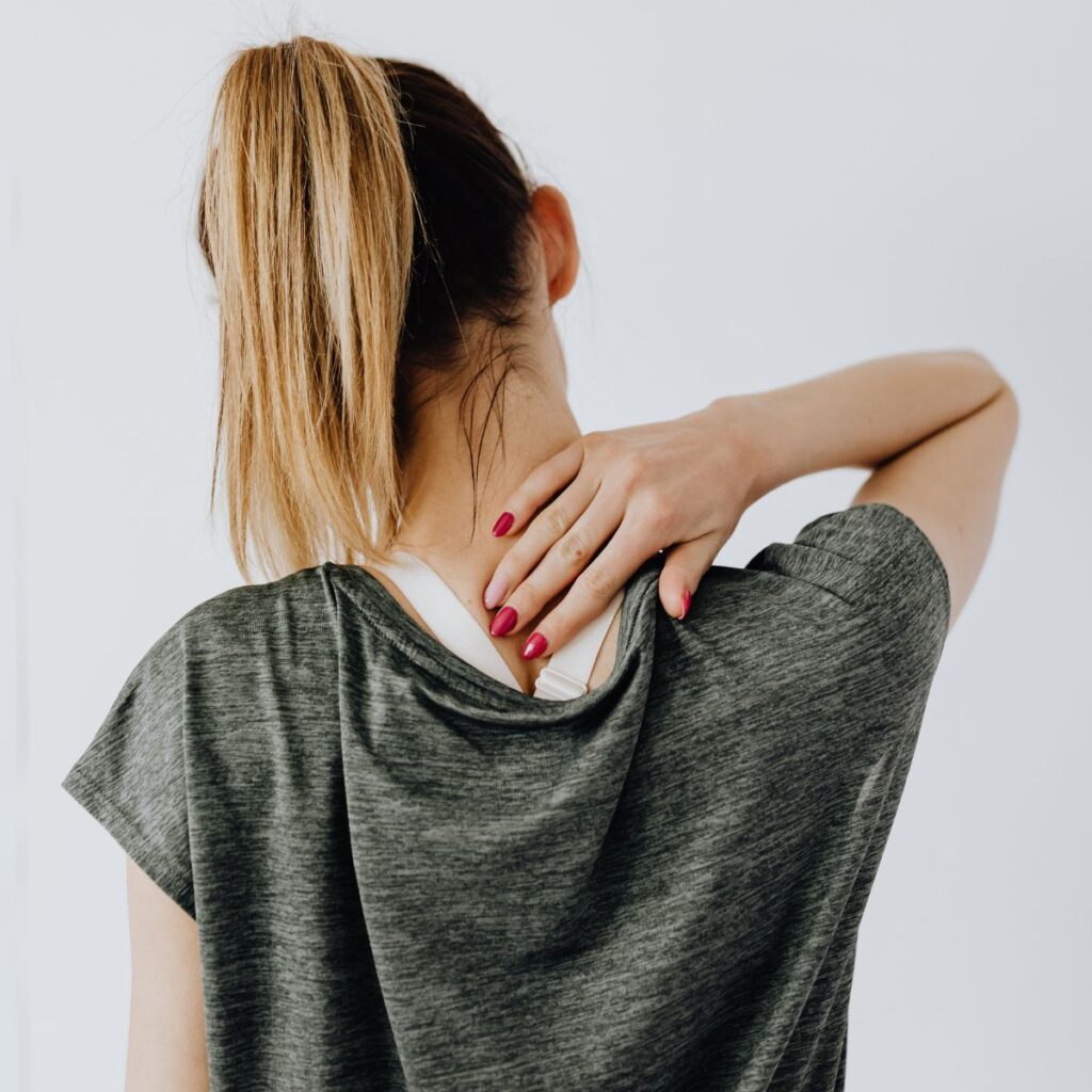 A woman with a ponytail, seen from behind, wearing a gray shirt, holds her neck with one hand as if experiencing pain or discomfort.