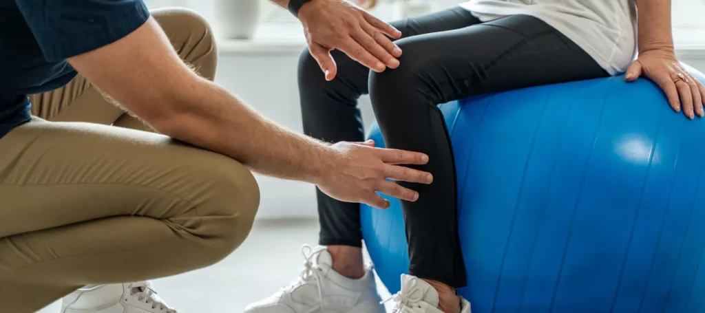 A person sits on a blue exercise ball while another person, possibly a physical therapist, supports and guides their leg during a rehabilitation or exercise session.