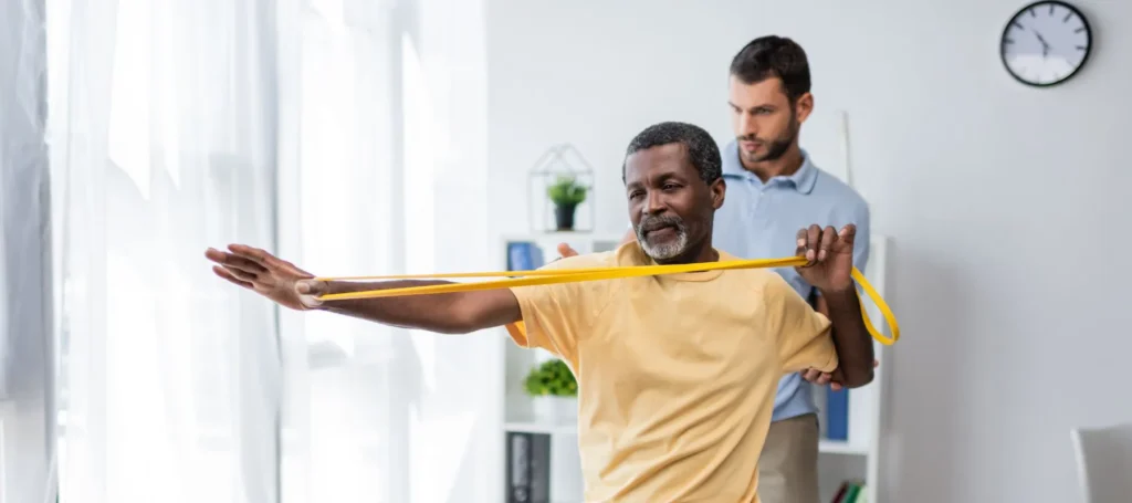 An older man in a yellow shirt stretches a yellow resistance band with one arm extended, while a therapist watches and guides him in a bright room with white curtains and a clock on the wall.