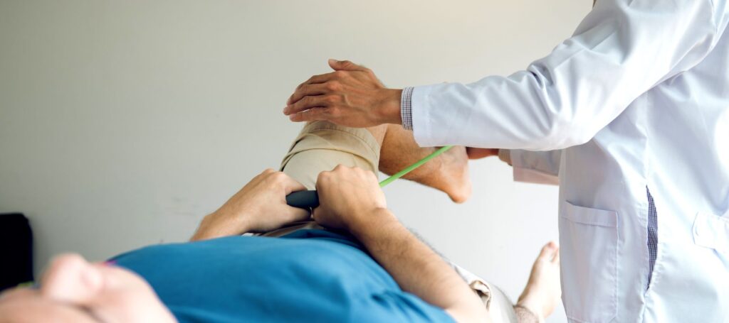A medical professional in a white coat measures the thigh of a person lying down, whose leg is wrapped in a bandage. The patient appears to be undergoing a physical examination or rehabilitation.