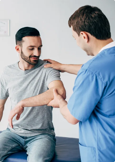 A healthcare professional in blue scrubs examines a seated mans arm, gently testing his elbow joint while the man looks down at his arm and smiles.