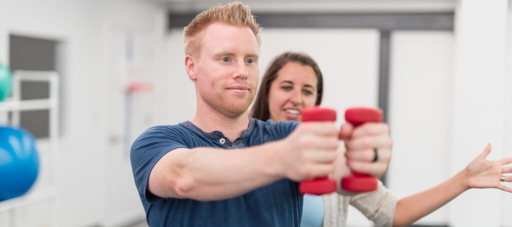 A man in a blue shirt lifts red dumbbells with both hands extended forward, while a woman behind him gestures supportively, possibly guiding him through an exercise.