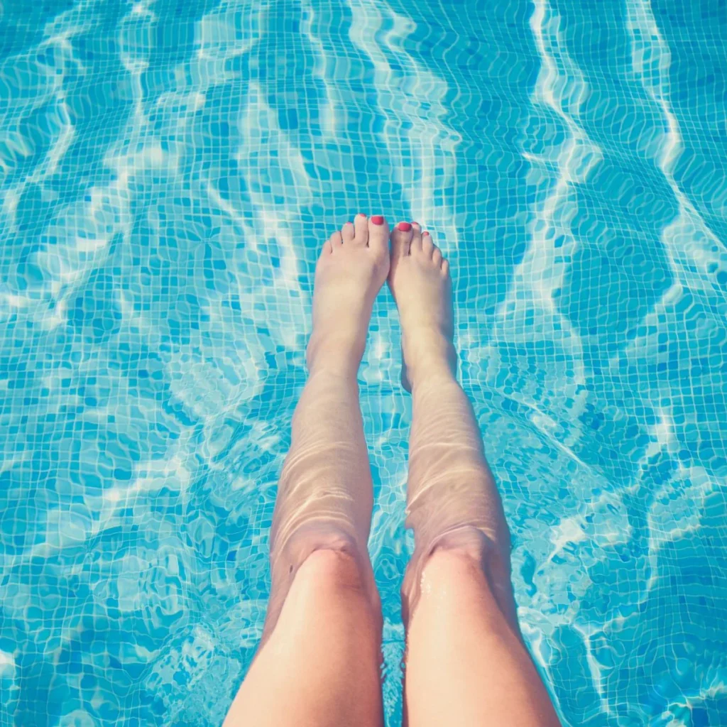 A person’s legs and feet are partially submerged in clear, blue swimming pool water with rippling light reflections on the surface.