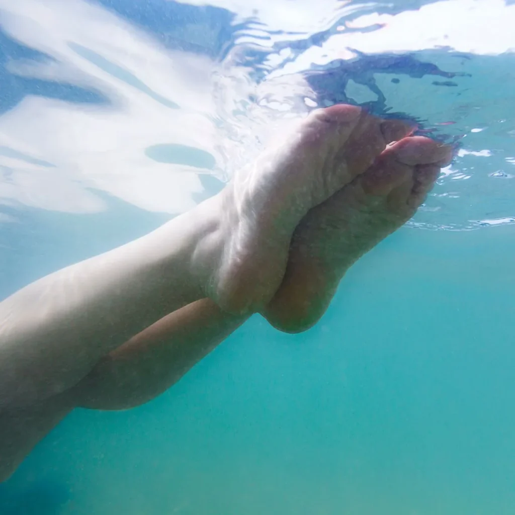 A close-up view of a persons bare feet and lower legs floating underwater, with sunlight filtering through the clear blue water above.