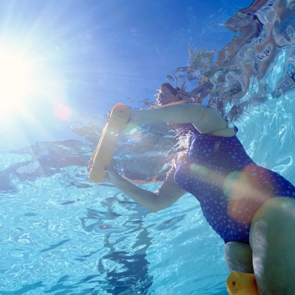 Underwater view of a person in a blue polka-dot swimsuit using a yellow pool noodle in a swimming pool, with sunlight streaming through the waters surface above.