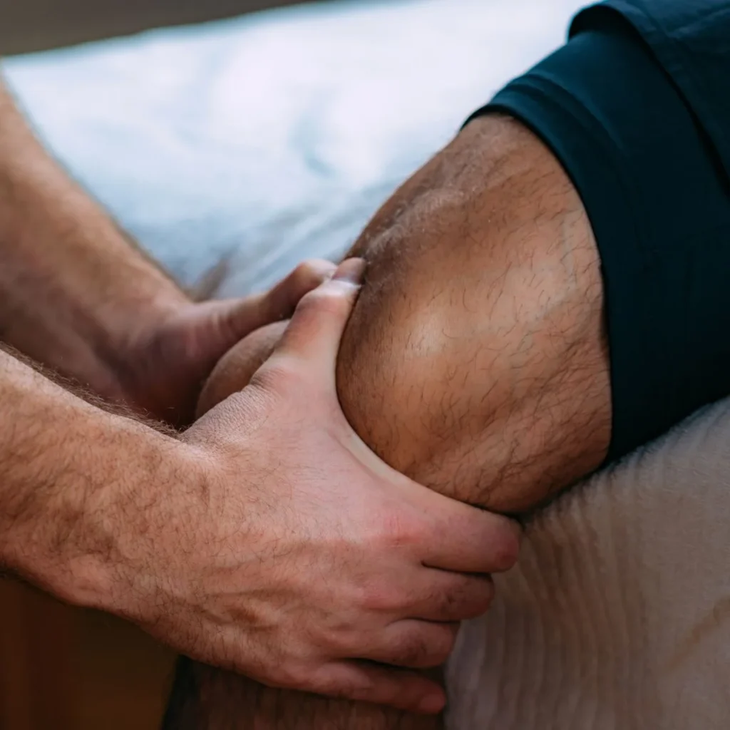 A close-up of a person’s hands gently holding and examining another person’s knee, possibly during a physical therapy or medical assessment. The person being examined is wearing dark shorts.