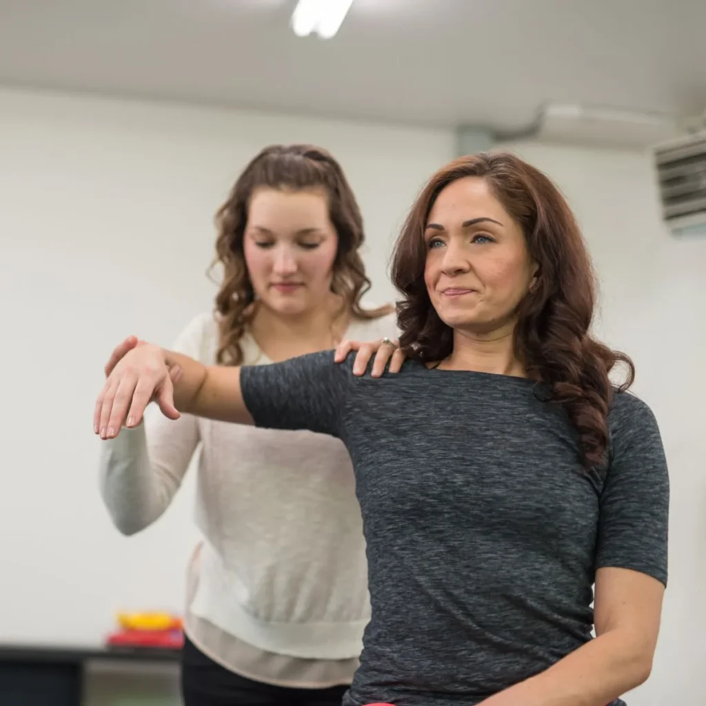 A woman stands with her arm extended while another woman, possibly a physical therapist, supports her back and arm, guiding her through a physical assessment or exercise in a bright indoor setting.