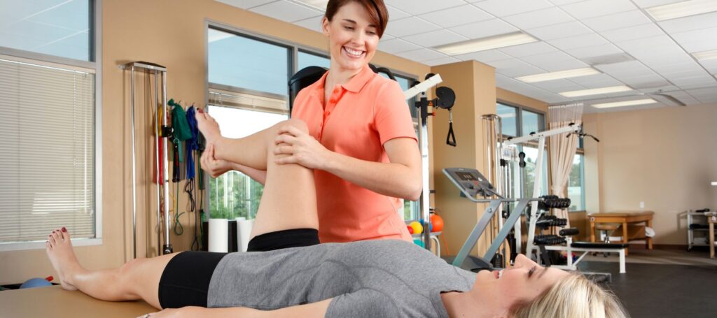 A physical therapist smiles while helping a patient stretch her leg on an exam table in a bright rehabilitation clinic equipped with exercise machines and therapy tools.
