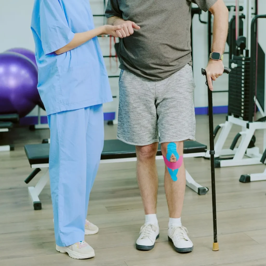 A person in athletic shorts with colorful kinesiology tape on their knee uses a cane for support. A healthcare professional in blue scrubs stands beside them, offering assistance in a gym or rehabilitation setting.