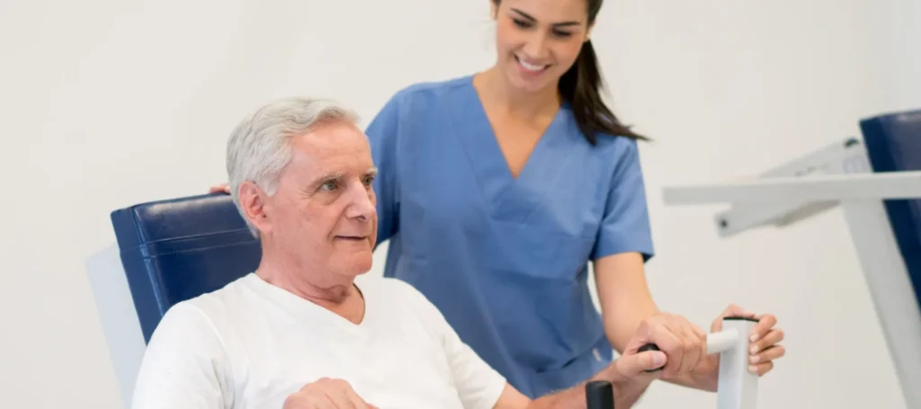An older man uses exercise equipment while a smiling female healthcare worker in blue scrubs assists him in a bright, clinical setting.