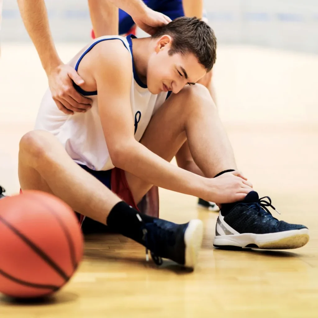 A young basketball player sits on the court, holding his ankle in pain while a teammate supports him. A basketball lies nearby on the wooden floor.